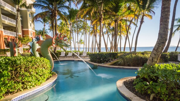 A swimming pool with a decorative sea creature sculpture, surrounded by palm trees and a hotel building.