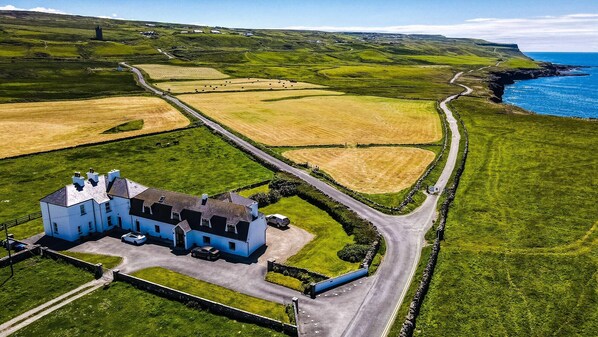 A large, white, castle-like building with a driveway and parked cars, surrounded by green fields and a coastal view.
