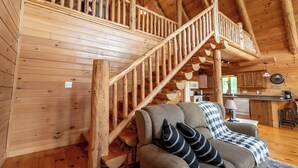 Ein Holzhütten-Interieur mit einer Treppe, die zu einer oberen Ebene führt, einer Couch mit kariertem Überwurf und einem Küchenbereich.