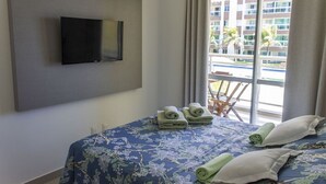 A hotel room with a bed, a flat-screen TV mounted on the wall, and a view of a building and palm trees outside. - Beachfront apartment in Praia do Futuro with full amenities. (Antonio Diogo)