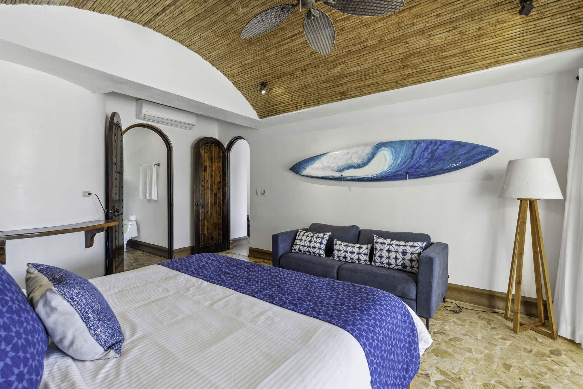 Ein Schlafzimmer mit zwei Betten, einer Holzbalkendecke, einem blau-weißen Sofabezug und einer Surfbrettausschnitt-Dekoration, sowie einer Lampe.