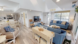 A spacious living area with a sofa, a coffee table, and a TV. There is a kitchen area with white cabinets and a dining table with chairs. - Great Multi-Family or Group Getaway, Walk To The Tybee Pier (Tybee Island)