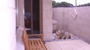 Un petit espace extérieur aménagé en patio, avec un banc en bois, un mur en béton et une porte donnant sur une entrée abritée.
