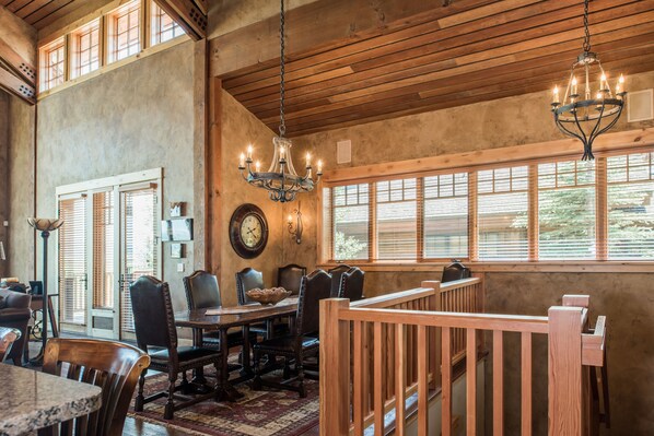A dining area with wooden furniture, a chandelier, and large windows with blinds.