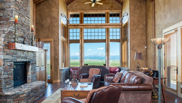A spacious living room with a stone fireplace, leather furniture, large windows, and a high ceiling with wooden beams. - Luxury home with Stunning 4 Peak Views (Tetonia)