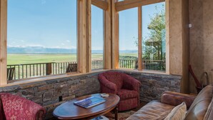 A room with a stone wall, a red armchair, a leather couch, and large windows offering a view of a green field and mountains. - Luxury home with Stunning 4 Peak Views (Tetonia)