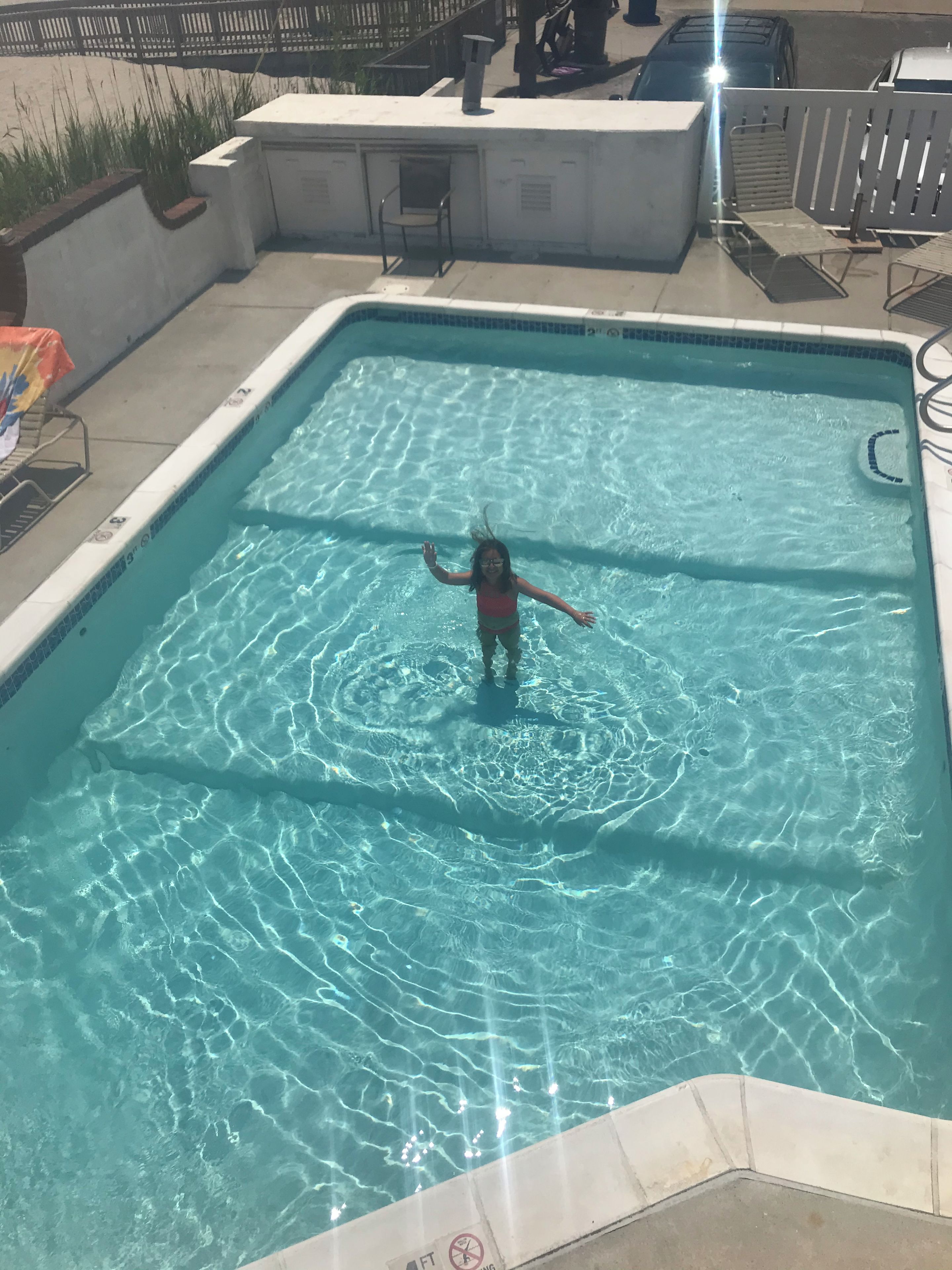 Ein Pool mit einem Kind im Wasser, umgeben von einer Sonnenterrasse mit Stühlen und einem Mülleimer.