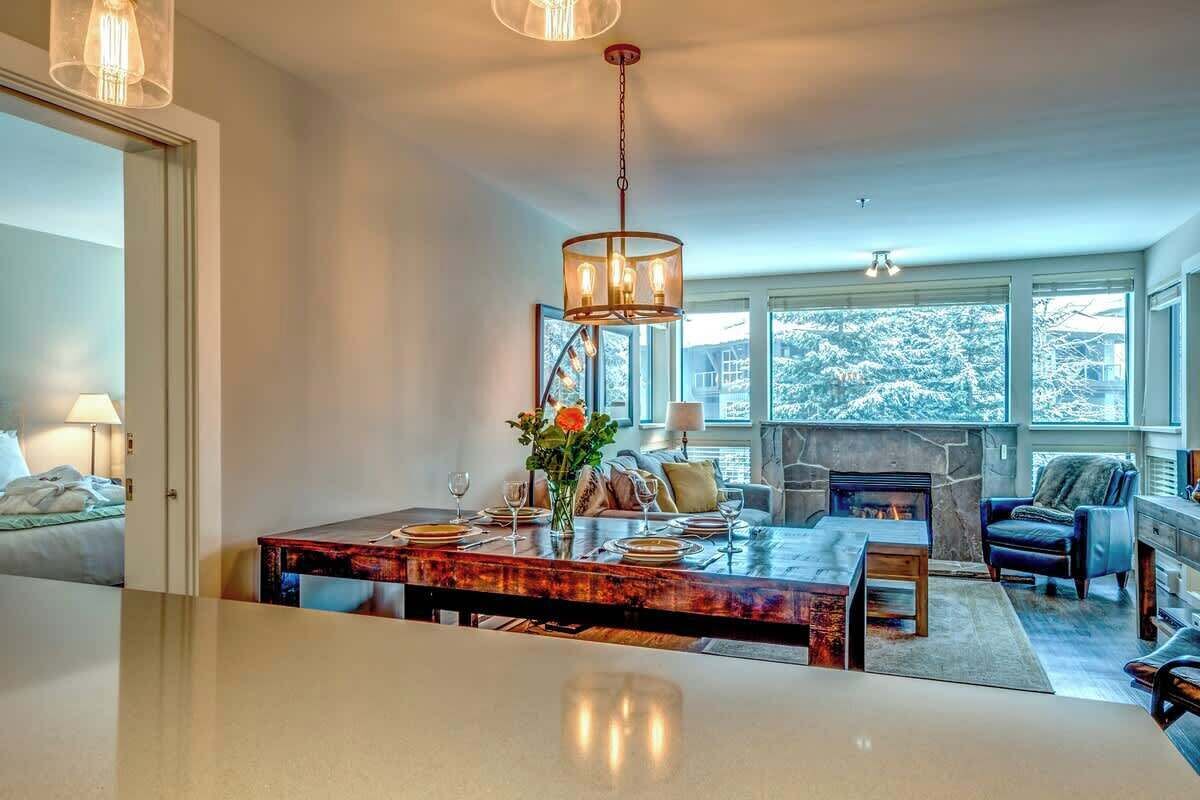 A modern dining area with a central table, a fireplace, and large windows offering a view of snow-covered trees.