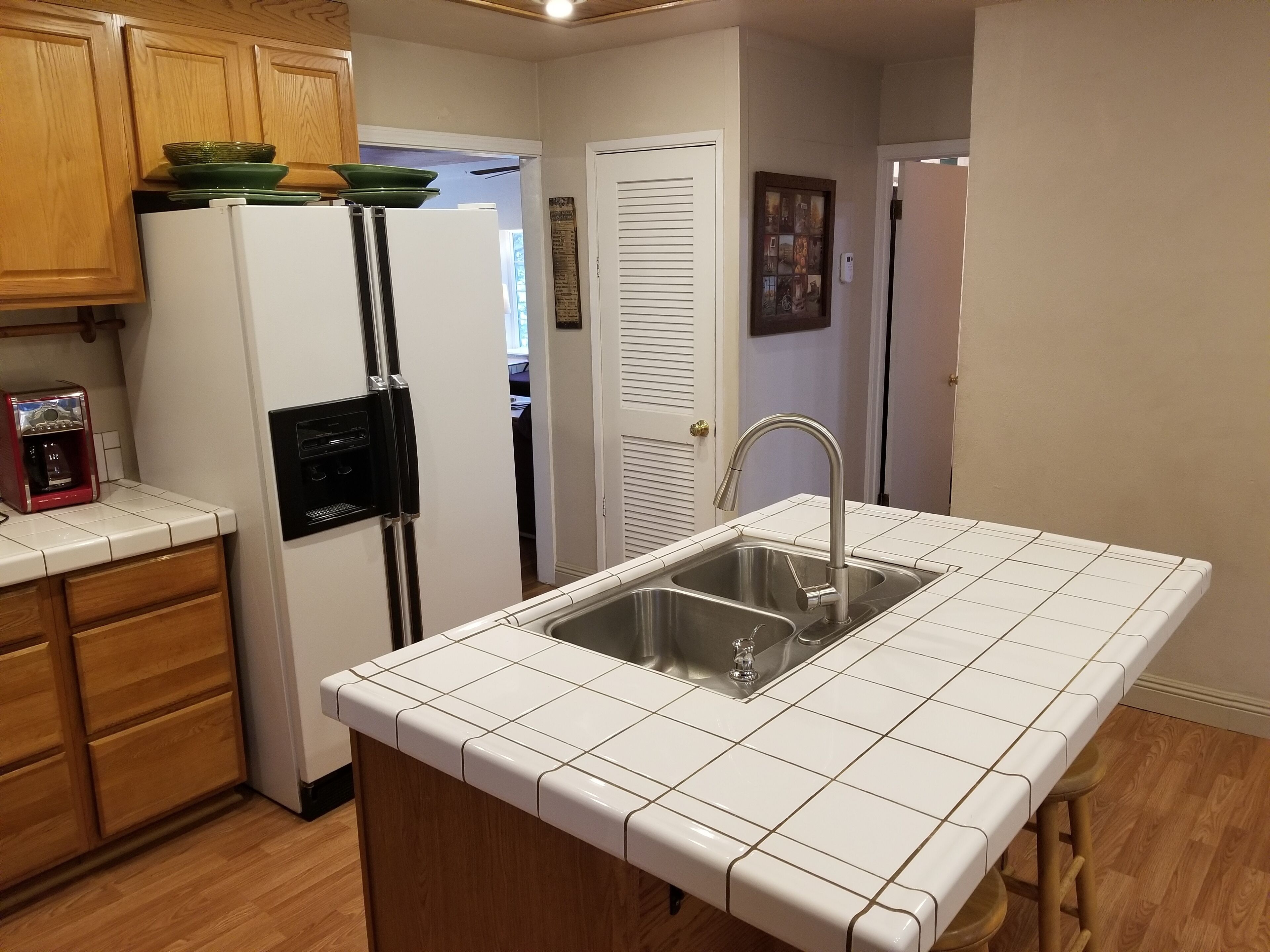 A kitchen with white tiled countertops, a stainless steel sink, wooden cabinets, and a refrigerator with a built-in ice maker.