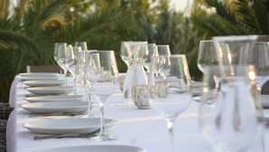 Mesa dispuesta para una comida, con platos, vasos y un centro de mesa, al aire libre con palmeras al fondo.