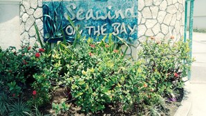 A sign with the text "Seaside on the Bay" against a stone wall, surrounded by various plants and flowers. - 24 gated security studio apartment,  Seawind on the bay Freeport Mobay (Montego Bay)