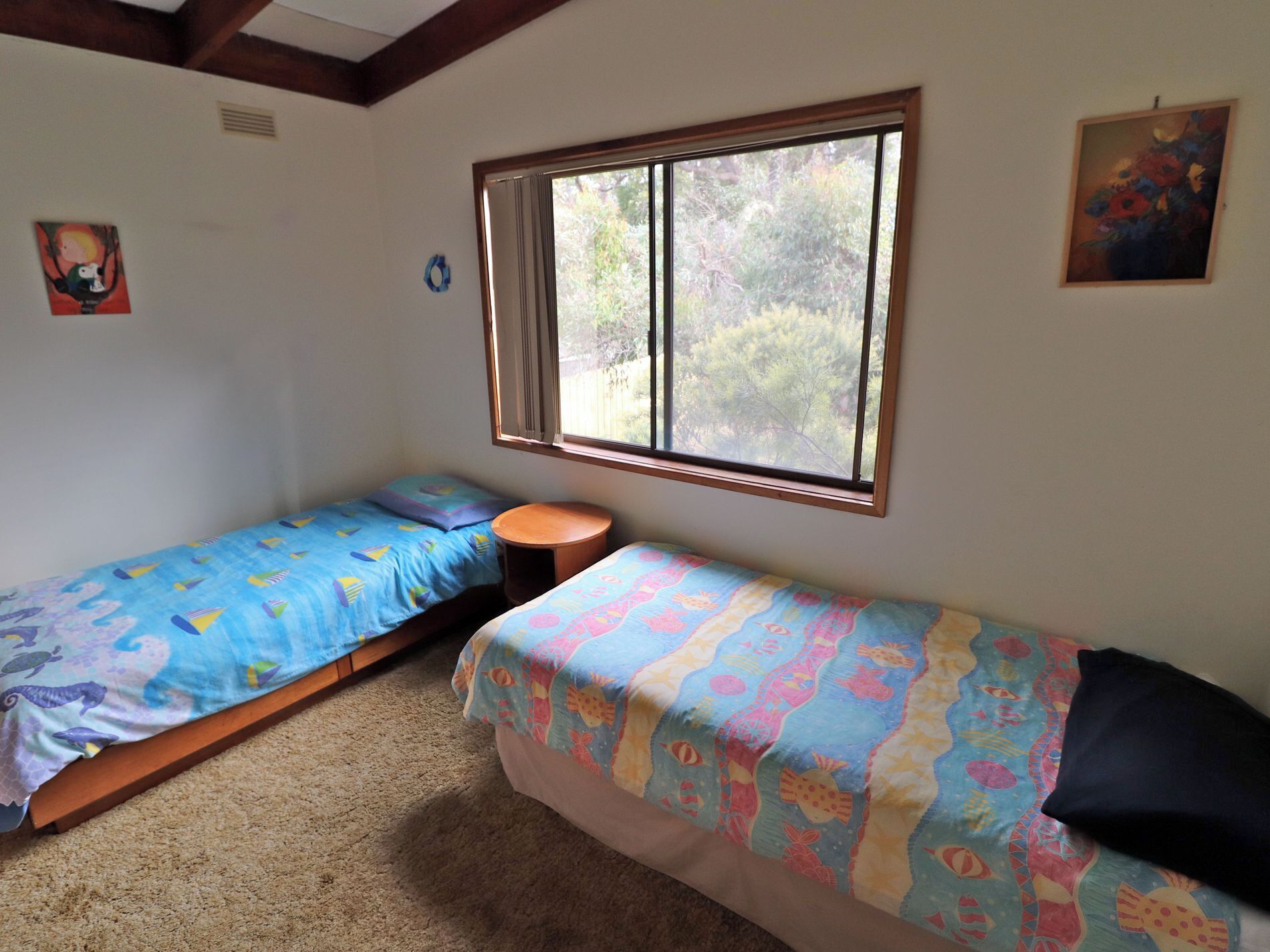 A small, cozy room with two single beds, a round wooden table, and a window providing a view of trees.