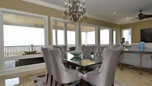 A dining area with a glass-top table surrounded by upholstered chairs, a chandelier, and a view of the outdoors through large windows. - Luxurious Oceanfront Home: Pool, SPA, & Movie Theater (Murrells Inlet)