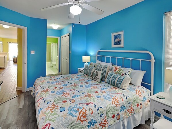 Un dormitorio con pared azul, ventilador de techo blanco y cama con colcha estampada, además de una mesita de noche con lámpara.