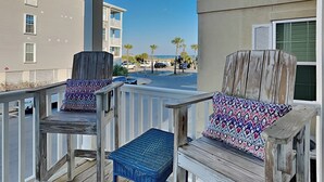 A balcony with two wicker chairs and a small table, overlooking a street with palm trees and a building in the background.