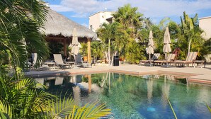 A clear swimming pool surrounded by lounge chairs and umbrellas, with palm trees and a thatched-roof building in the background.