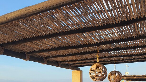 A covered outdoor seating area with a sofa and table, a thatched roof, and a view of the landscape.