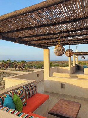 A covered outdoor seating area with a sofa and table, a thatched roof, and a view of the landscape.