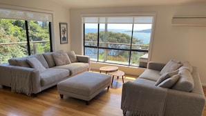 Ein modernes Wohnzimmer mit zwei grauen Sofas, einem kleinen runden Tisch und großen Fenstern mit Blick auf das Meer und Grünflächen.