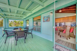 A covered outdoor area with a dining table and chairs, a kitchen area with wicker furniture, and a view of the outdoors through large windows.