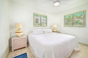 A bedroom with a bed, two nightstands, two lamps, a ceiling fan, and two windows with shutters.