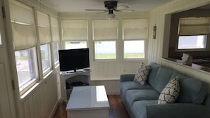 Ein Wohnzimmer mit Fernseher auf einem Schrank, einem weißen Couchtisch, einem blauen Sofa mit gemusterten Kissen und einer Sitzbank an einem Fenster.