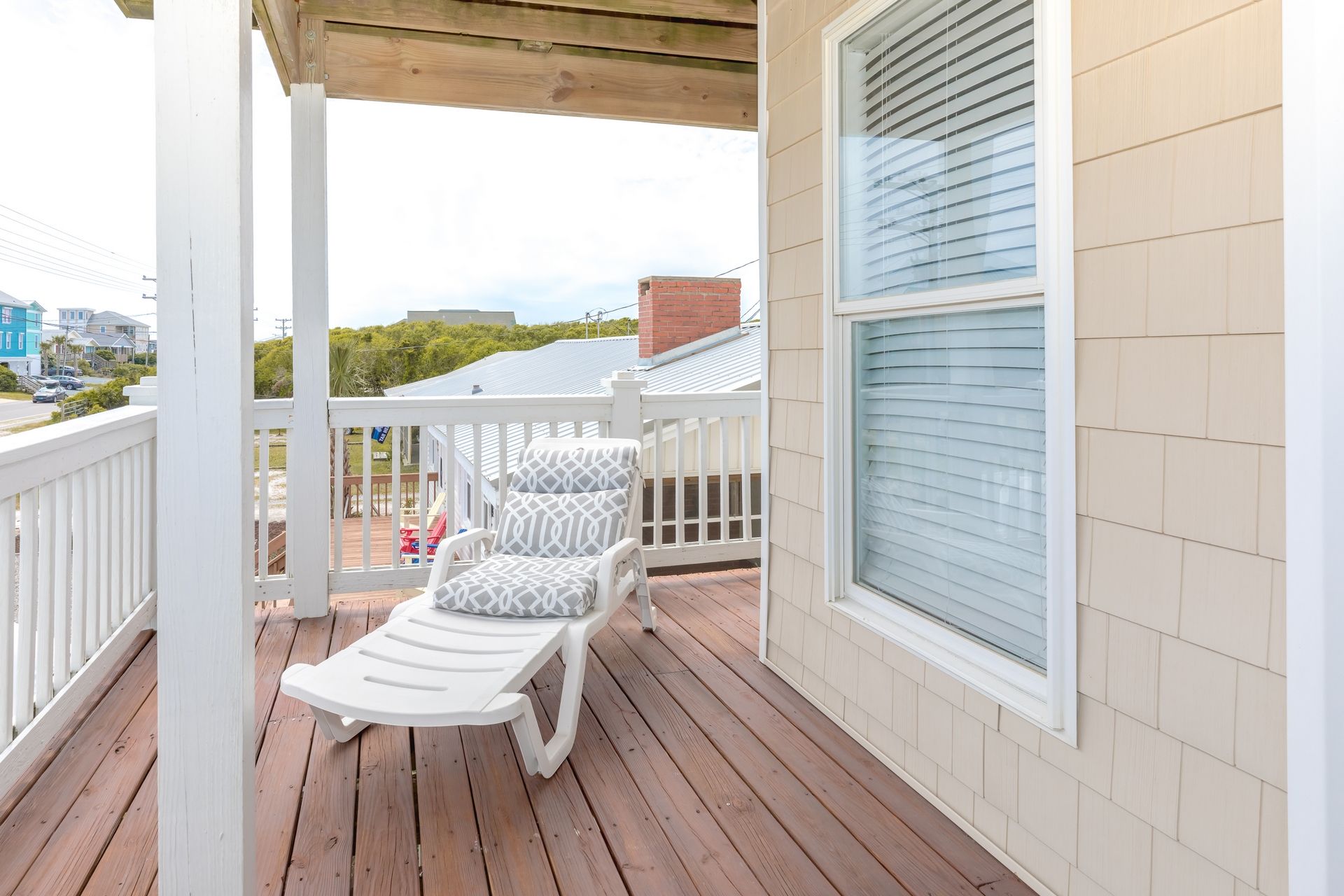 A covered outdoor seating area with a white railing, a single lounge chair, and a window with blinds.