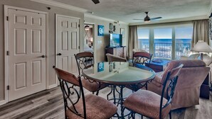 A dining area with a round glass table and metal chairs, a ceiling fan, and a view of the ocean through large windows. - Gulf Front Sea Oats 303 with Rocker Recliner, Comfy beds and Beach Service!  (Fort Walton Beach)