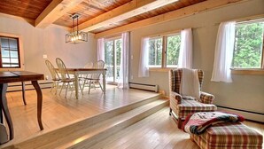 Una habitación amplia con suelo de madera, una mesa de comedor con sillas, un sillón a cuadros y una ventana con cortinas.