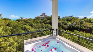 A balcony with a hot tub filled with water and rose petals, surrounded by lush greenery and a view of a building with a red roof.