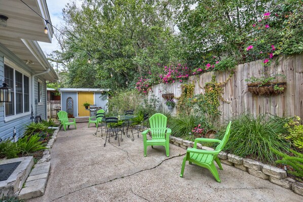 A patio with green chairs and a small table, surrounded by plants and a wooden fence.