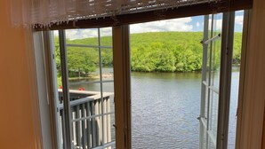 A window with a view of a lake, a wooden deck, and a grassy hill.