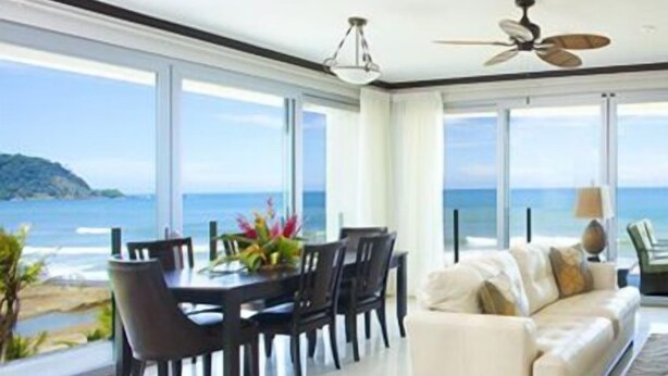 A modern living room with a white sofa, a dining table with chairs, and large windows offering a view of the ocean. - Manhattan hits Jaco 3br/3.5 ba Panoramic Ocean Views (Jaco)