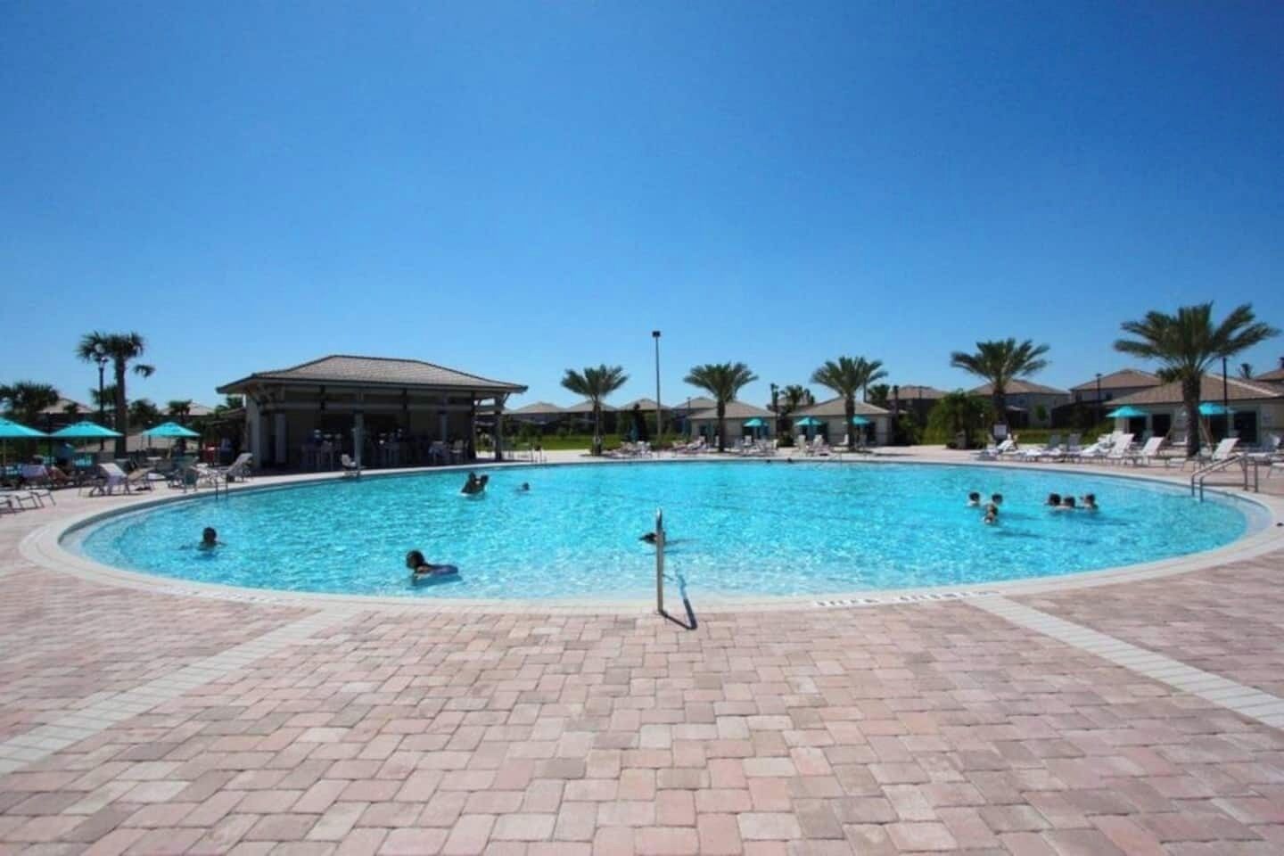 Una gran piscina al aire libre con gente nadando y relajándose. Palmeras y sombrillas se ven al fondo.
