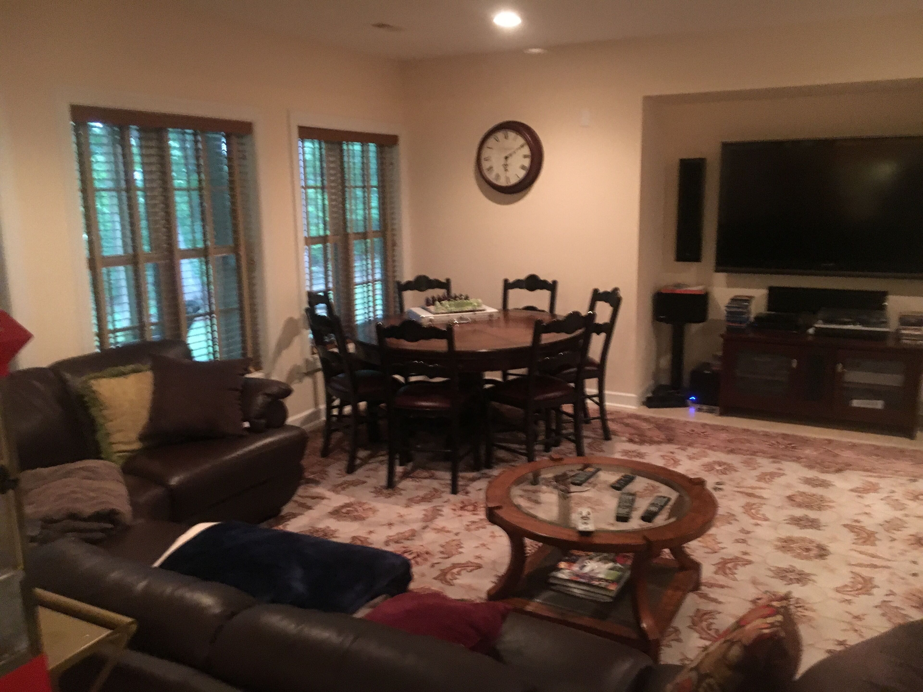 A living room with a leather sofa, a dining table with chairs, a coffee table, a TV, and a clock on the wall.