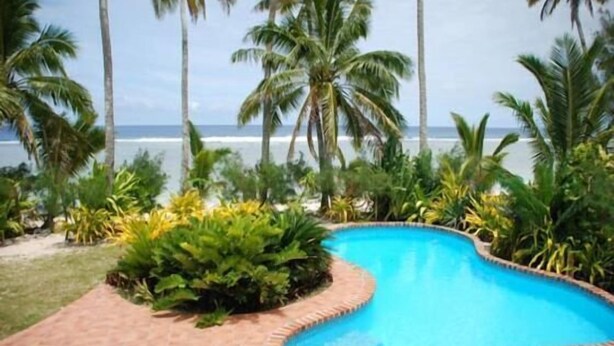 A swimming pool with a curved edge, surrounded by lush greenery and palm trees, with a view of the ocean in the background. - Heritage, Pool and AirCon (Arorangi)