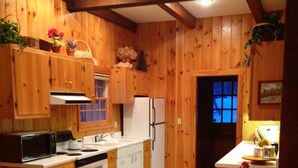 Cocina con paneles de madera, un refrigerador, estufa y microondas blancos. Hay armarios de madera, un fregadero y una pequeña isla con taburetes.