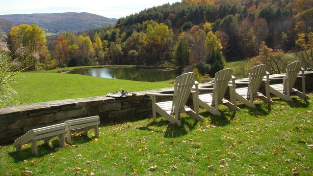Vista al lago con sillas Adirondack y un banco, rodeado de follaje otoñal y un muro de piedra.