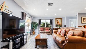 A modern living room with a flat-screen TV, a leather sofa, a wooden coffee table, and a large window with curtains. - 6bdr/4bath tropical paradise with heated pool and lanai, 1 mile from the beach (Naples)