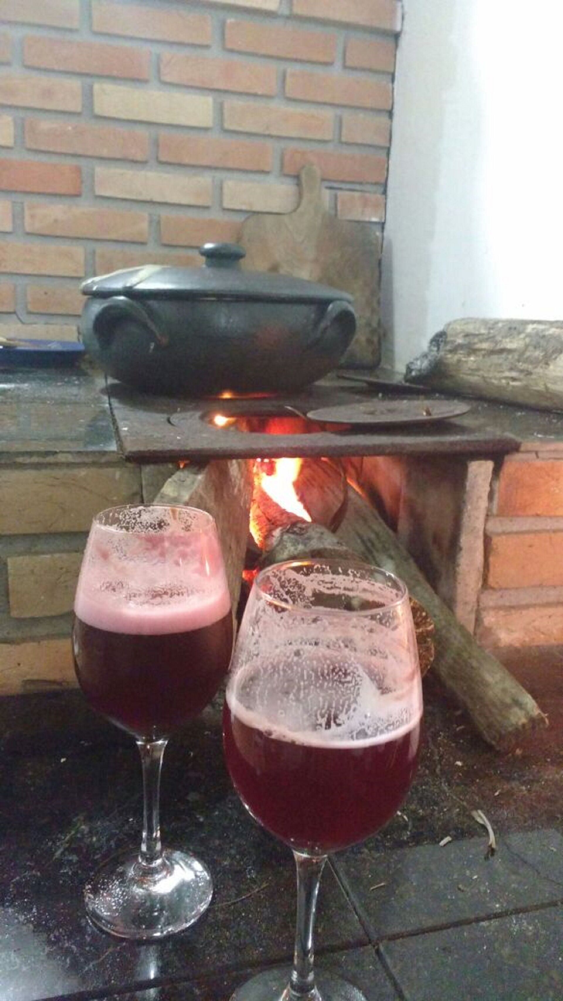 Two glasses of red wine in front of a brick fireplace with a pot on the mantel.