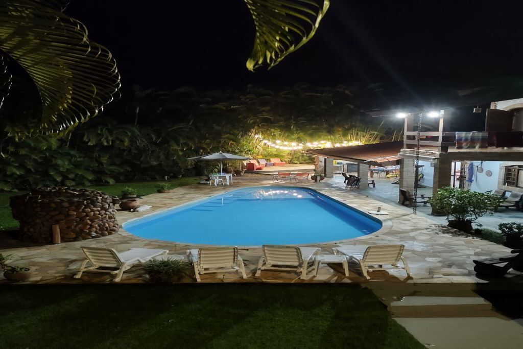 A swimming pool with lounge chairs, a stone patio, and a well-lit outdoor area with tables and umbrellas.