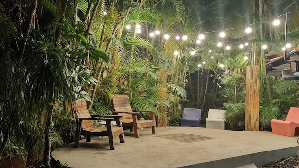 An outdoor seating area with wooden chairs and tables, surrounded by lush tropical plants and string lights.
