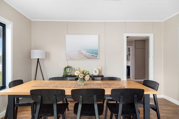 A dining area with a wooden table, black chairs, a vase with flowers, and a painting of a beach.
