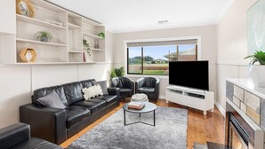 Een moderne woonkamer met een zwarte leren bank, een salontafel en een flatscreen televisie. Er zijn planken met decoratieve objecten en een open haard.