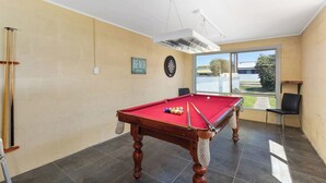 Een pooltafel met ballen en stokken, een dartbord, een bordje met de tekst 'STRAND DEZE KANT', en een uitzicht op een gebouw en groen buiten.