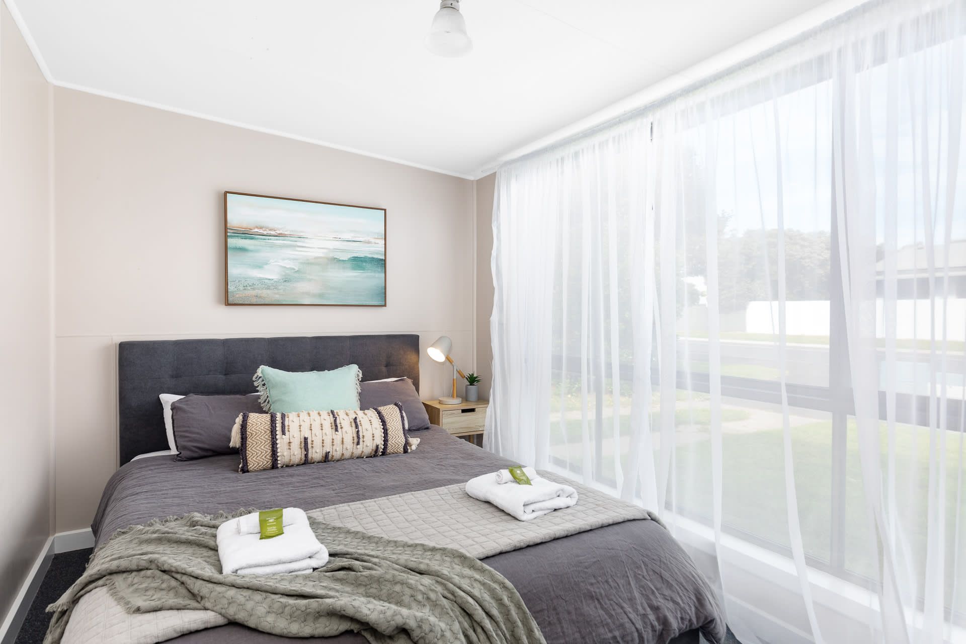 A bedroom with a bed, a bedside table, a lamp, and a window with sheer curtains.