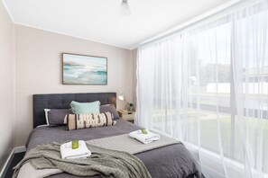 A modern bedroom with a large bed, a bedside table with a lamp, and a view of the outdoors through sheer curtains.