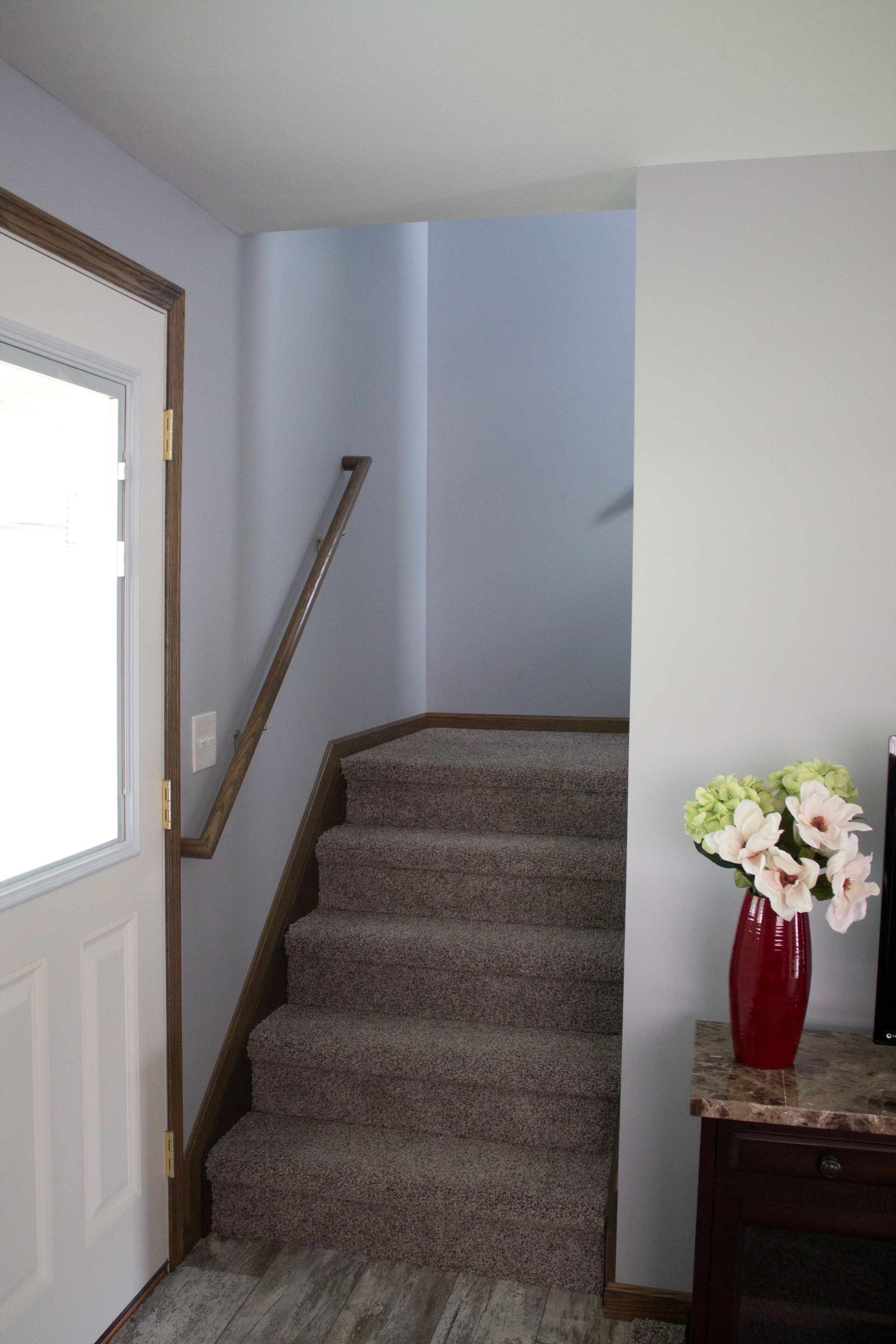 Una escalera con escalones alfombrados, una barandilla de madera y un jarrón rojo con flores sobre una mesa de madera.