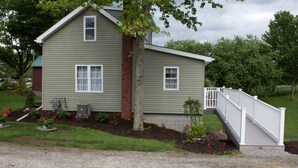 A two-story house with a brick chimney, a front porch with a white railing, and a small garden with flowers and shrubs. - Little house on the farm (Middlefield)