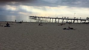 A beach with a pier extending into the water, people walking and relaxing on the sand, and a cloudy sky. - Hindmarsh House on College | Steps to Glenelg Beach, Cafes & Jetty Road (Glenelg)